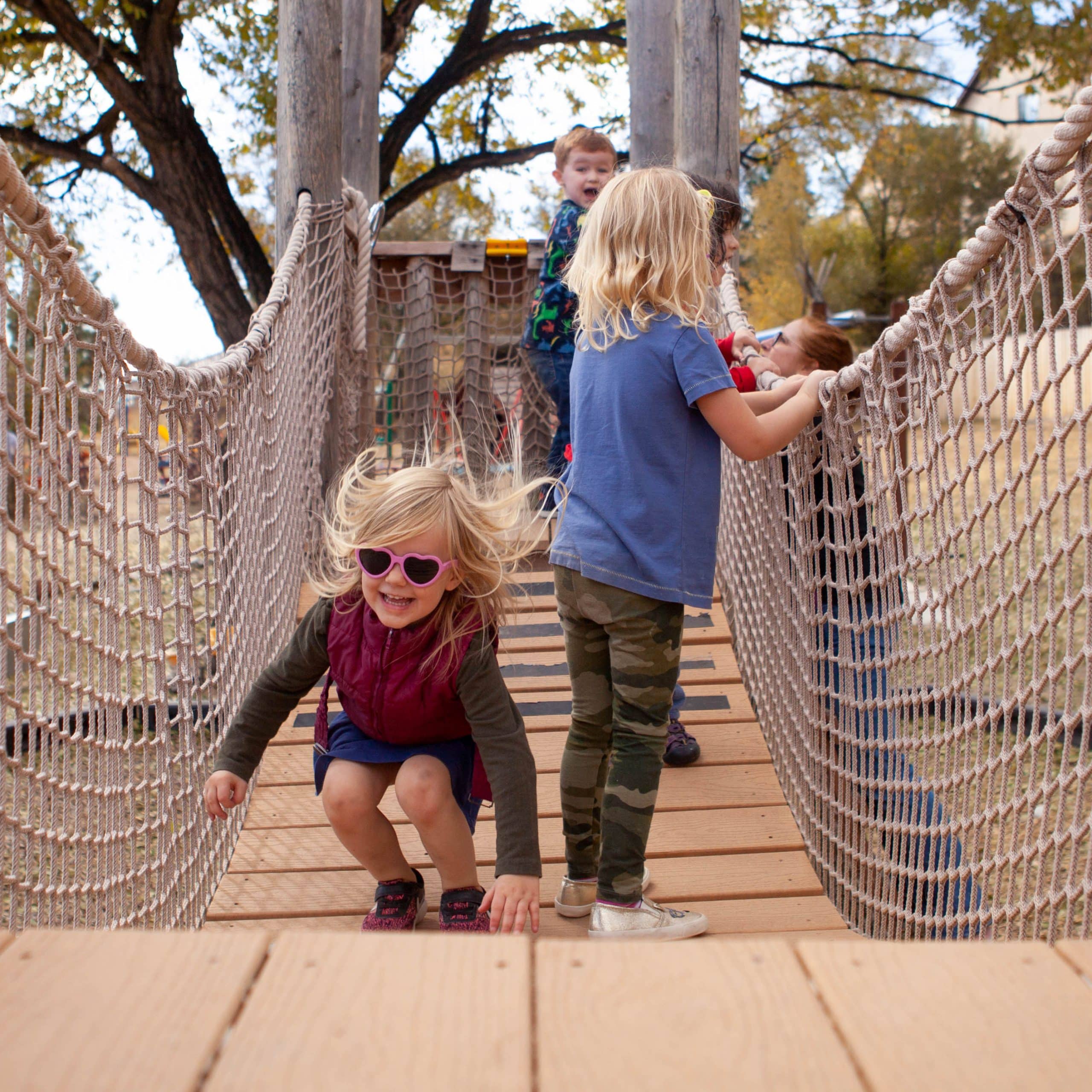 children walk across a wooden bridge with rope sides. One child with blond hair and sunglasses lands from a jump