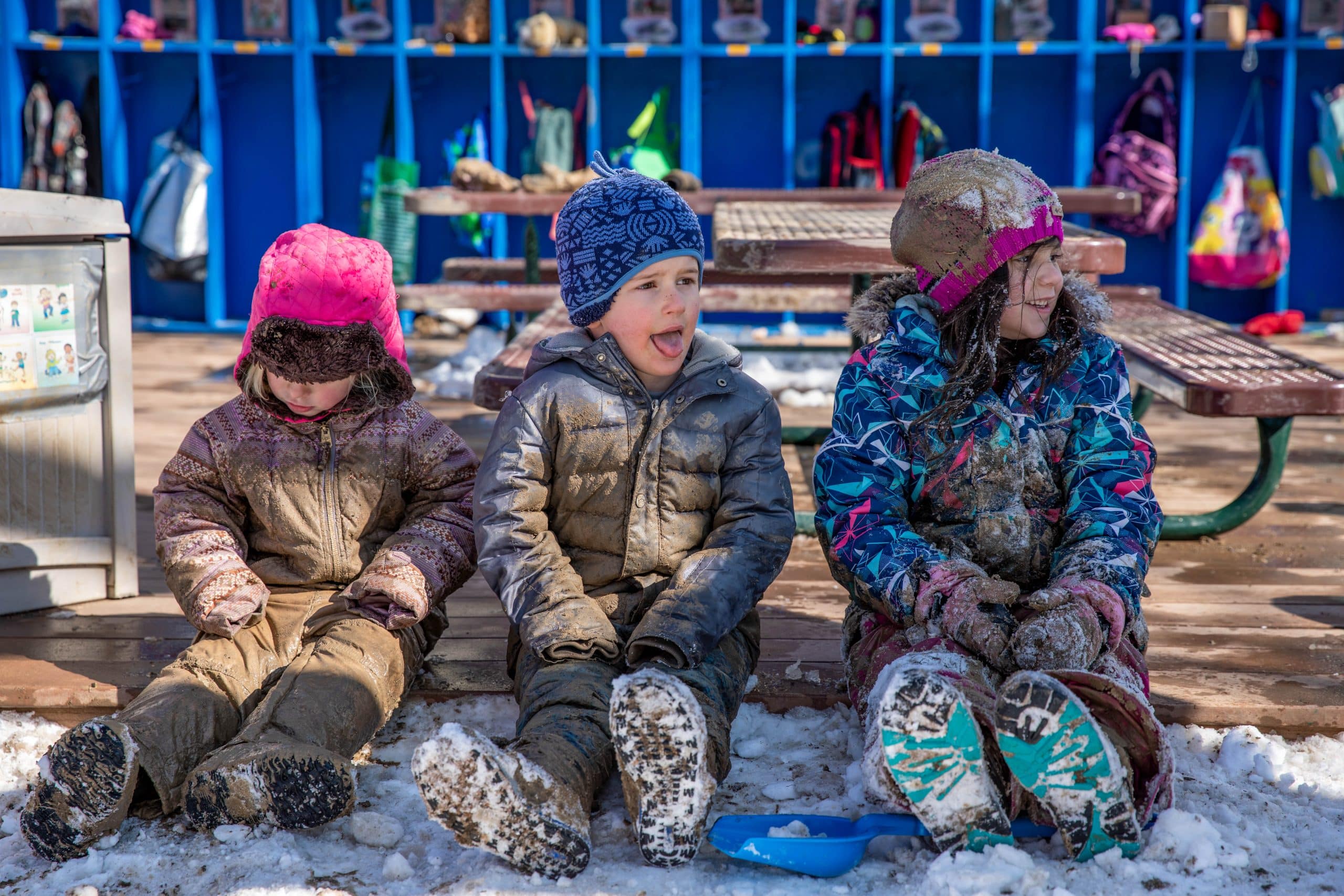 three children, wearing snow gear and covered in mud, sit on a deck in front of a row of blue cubbies