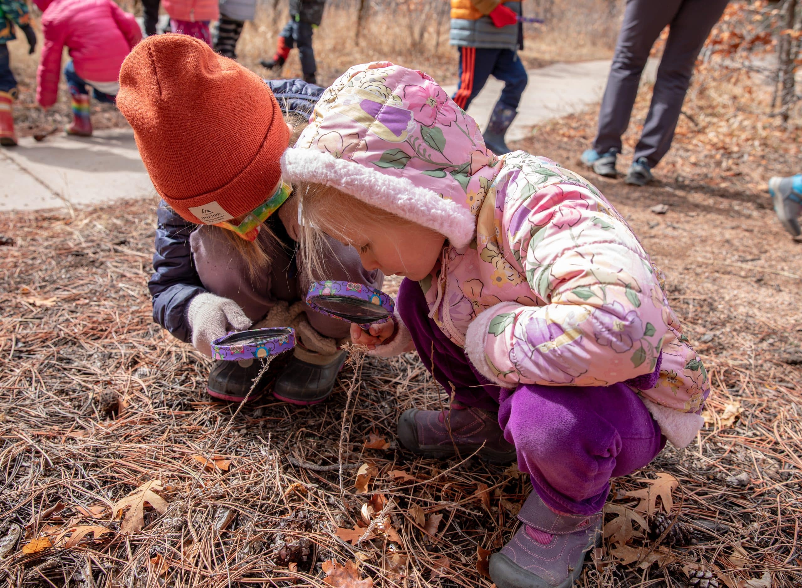 two children wearing winter coats look at plants through magnifying glasses