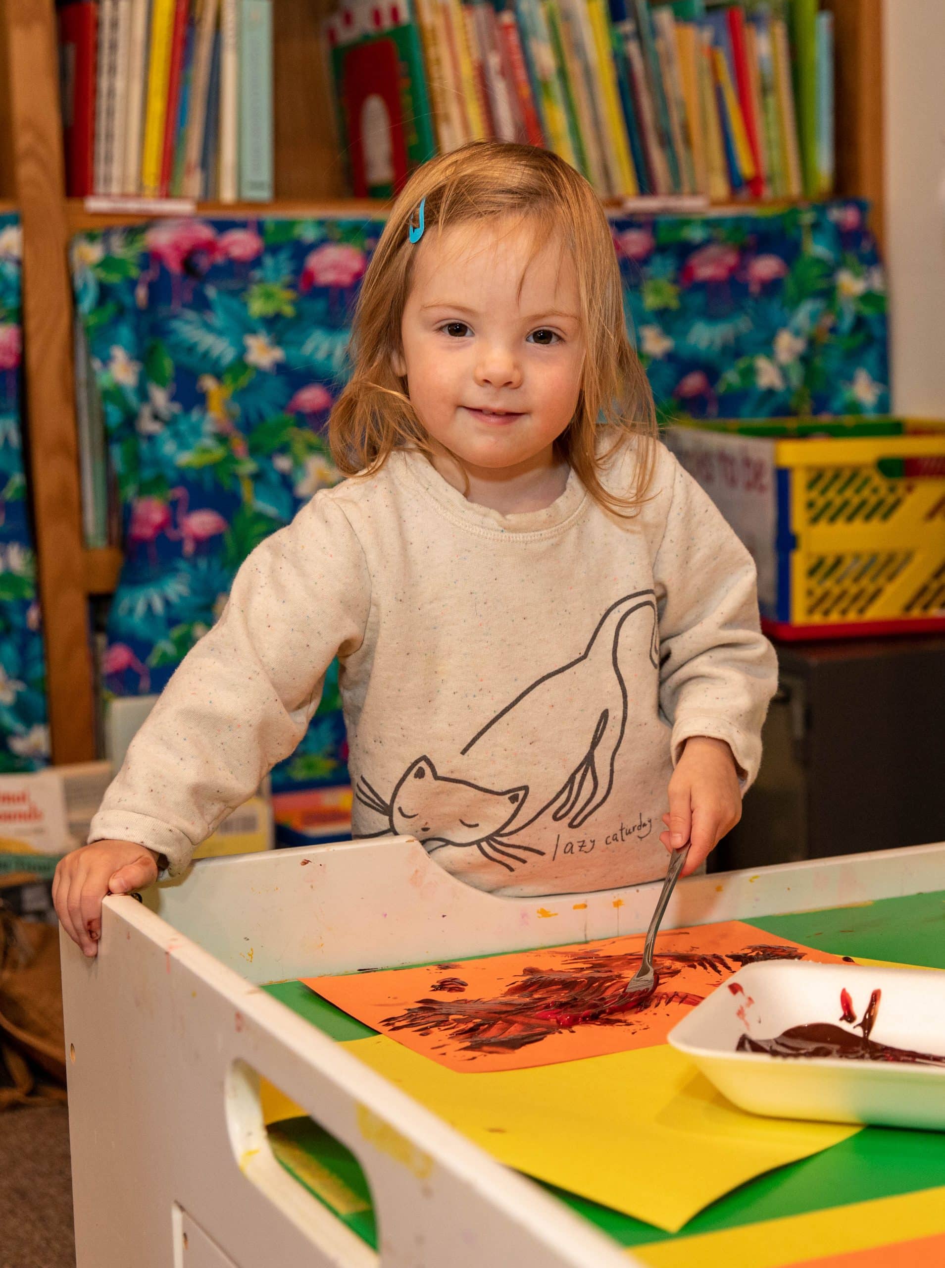 a child with long, blond hair painting with a fork onto brightly colored paper